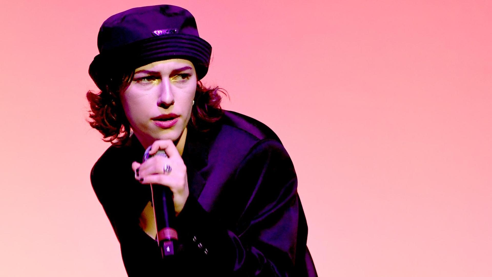 King Princess mit Mikro in der Hand vor einem rosa Hintergrund. King Princess mit Mikro in der Hand vor einem rosa Hintergrund.