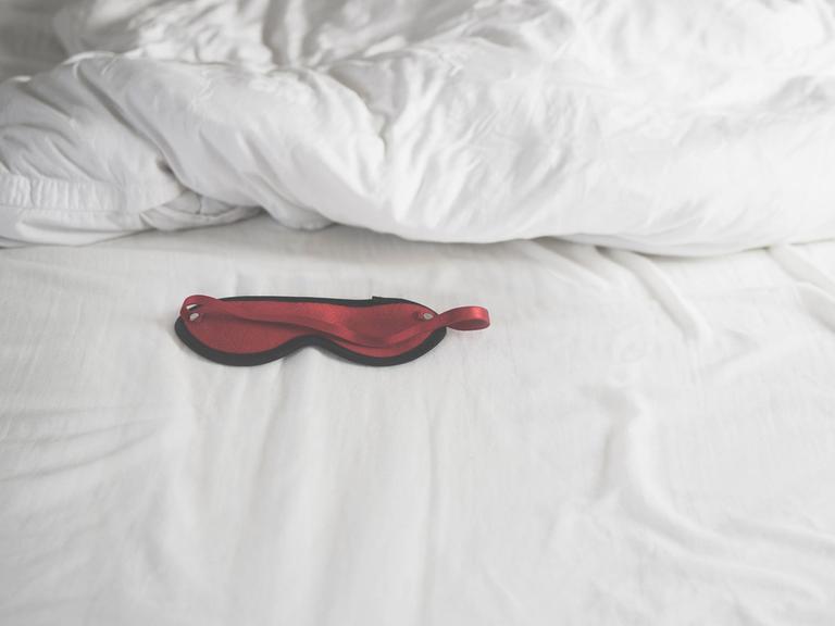 Eine rote, mit schwarzem Rand eingefasste Schlafmaske auf einem weißen Bettlaken. Dahinter ein weiß überzogenes Kopfkissen