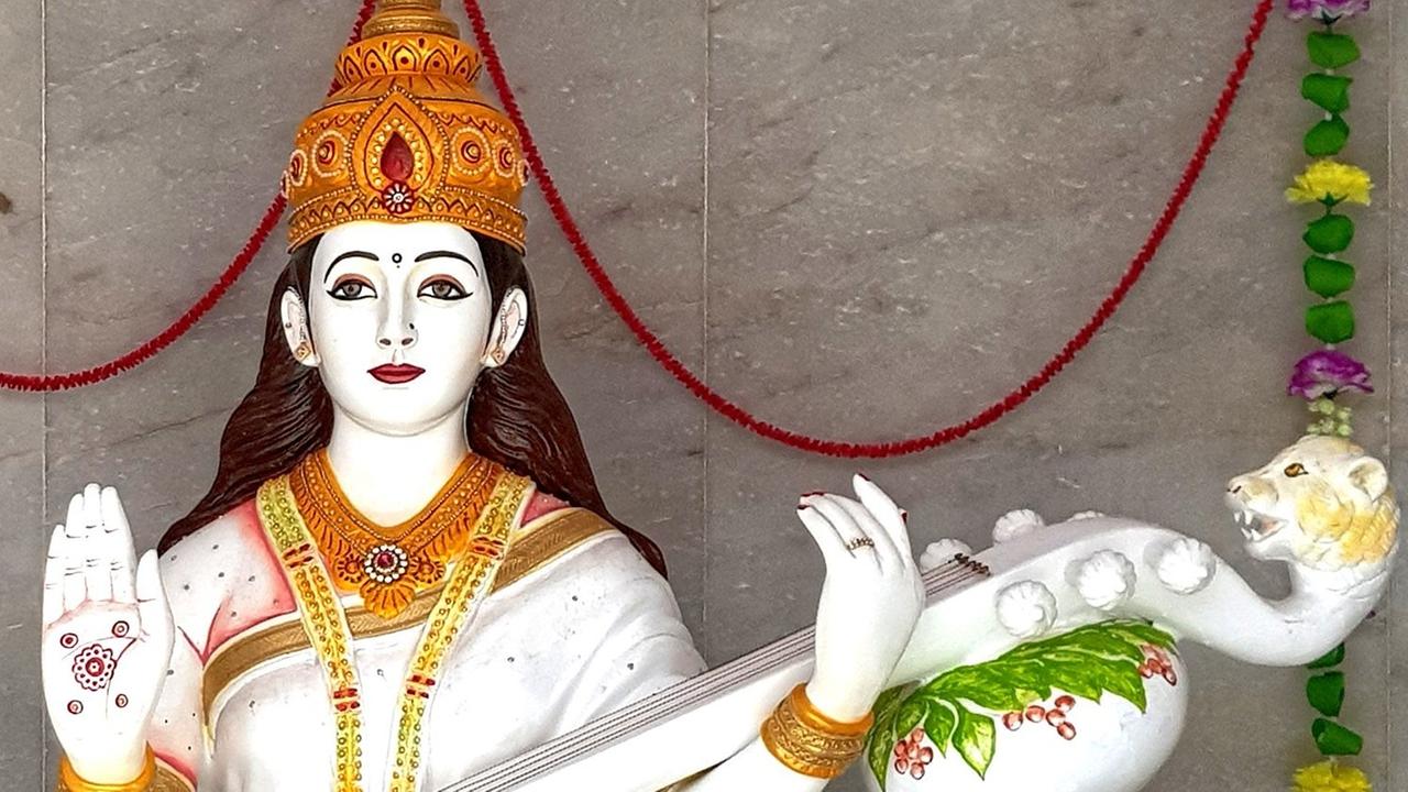 Skulptur der hinduistischen Gottheit Sarasvati, Göttin der Musik, mit goldener Krone und erhobener rechter Hand, in der anderen Hand hält sie ein Saiteninstrument mit Löwenkopf. Skulptur der hinduistischen Gottheit Sarasvati, Göttin der Musik, mit goldener Krone und erhobener rechter Hand, in der anderen Hand hält sie ein Saiteninstrument mit Löwenkopf.