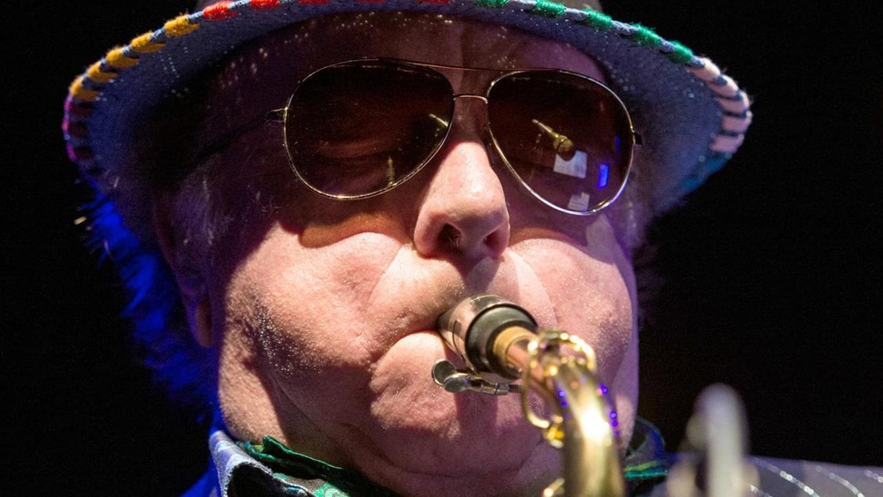 Das Bild zeigt Van Morrison mit Hut und Brille vor schwarzem Hintergrund. Er bläst in ein Saxofon.