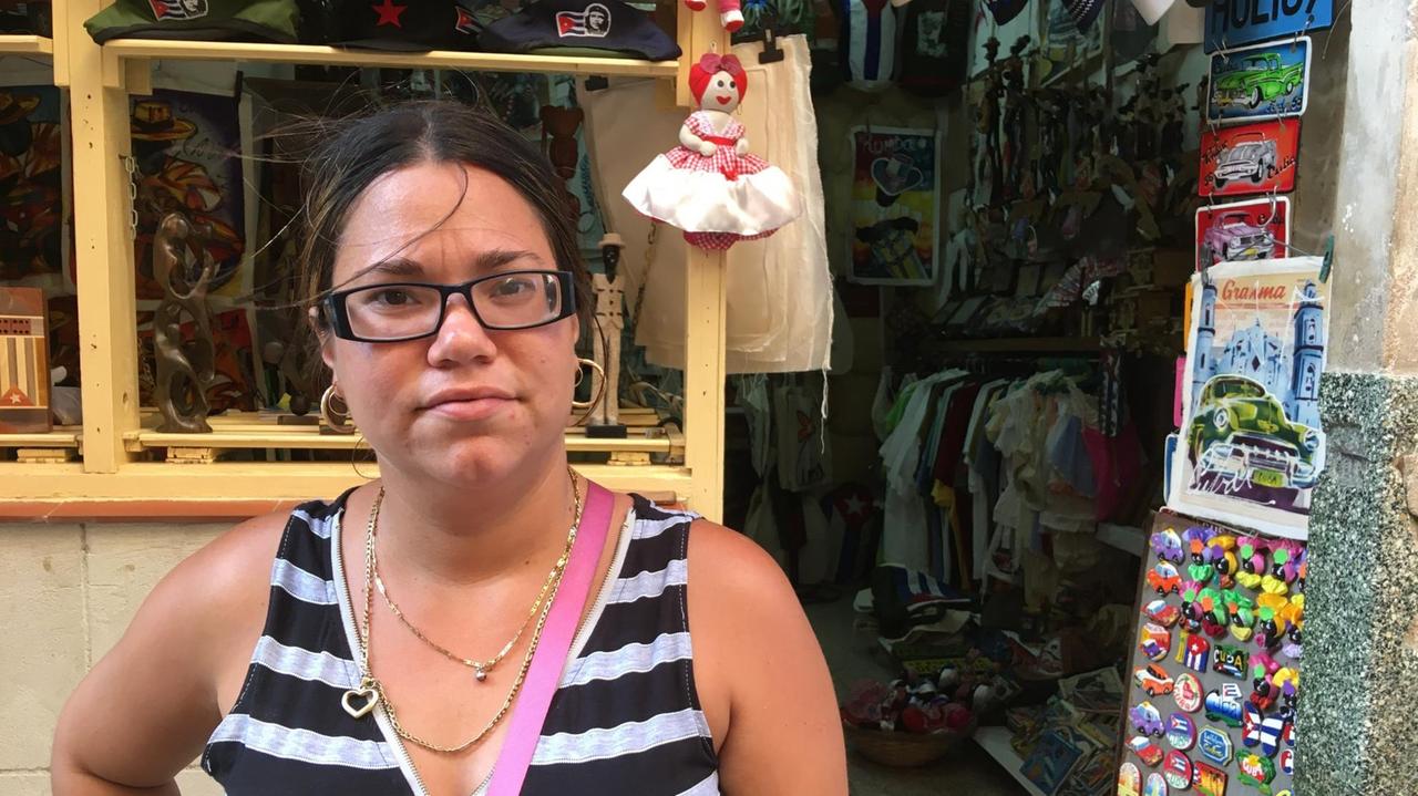 Eine junge Frau mit dunklem Zopf, Brille und Goldkettchen um den Hals steht vor ihrem Souvenirgeschäft. Eine junge Frau mit dunklem Zopf, Brille und Goldkettchen um den Hals steht vor ihrem Souvenirgeschäft.