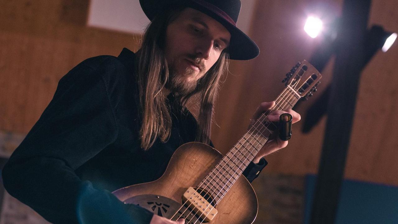 Ein Mann mit Hut und langen Haaren hält eine Gitarre in den Händen. Im Hintergrund strahlt ein Scheinwerfer. Ein Mann mit Hut und langen Haaren hält eine Gitarre in den Händen. Im Hintergrund strahlt ein Scheinwerfer.