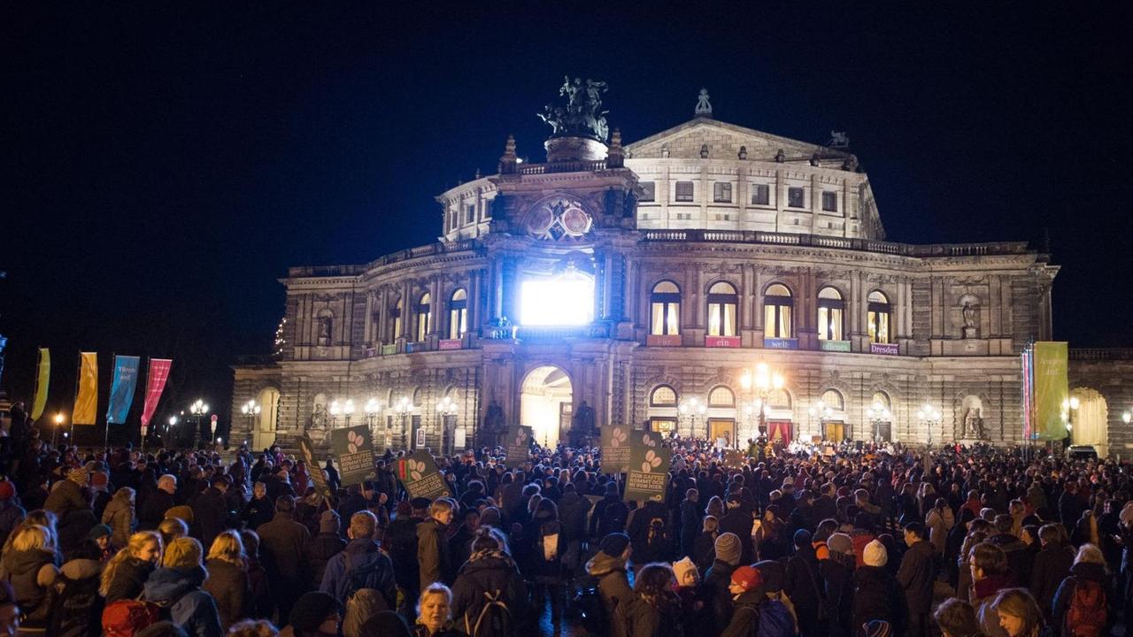 Zu sehen ist eine Menschenmenge, es ist Abend, im Hintergrund sieht man die Semperoper, die erleuchtet ist. Zu sehen ist eine Menschenmenge, es ist Abend, im Hintergrund sieht man die Semperoper, die erleuchtet ist.