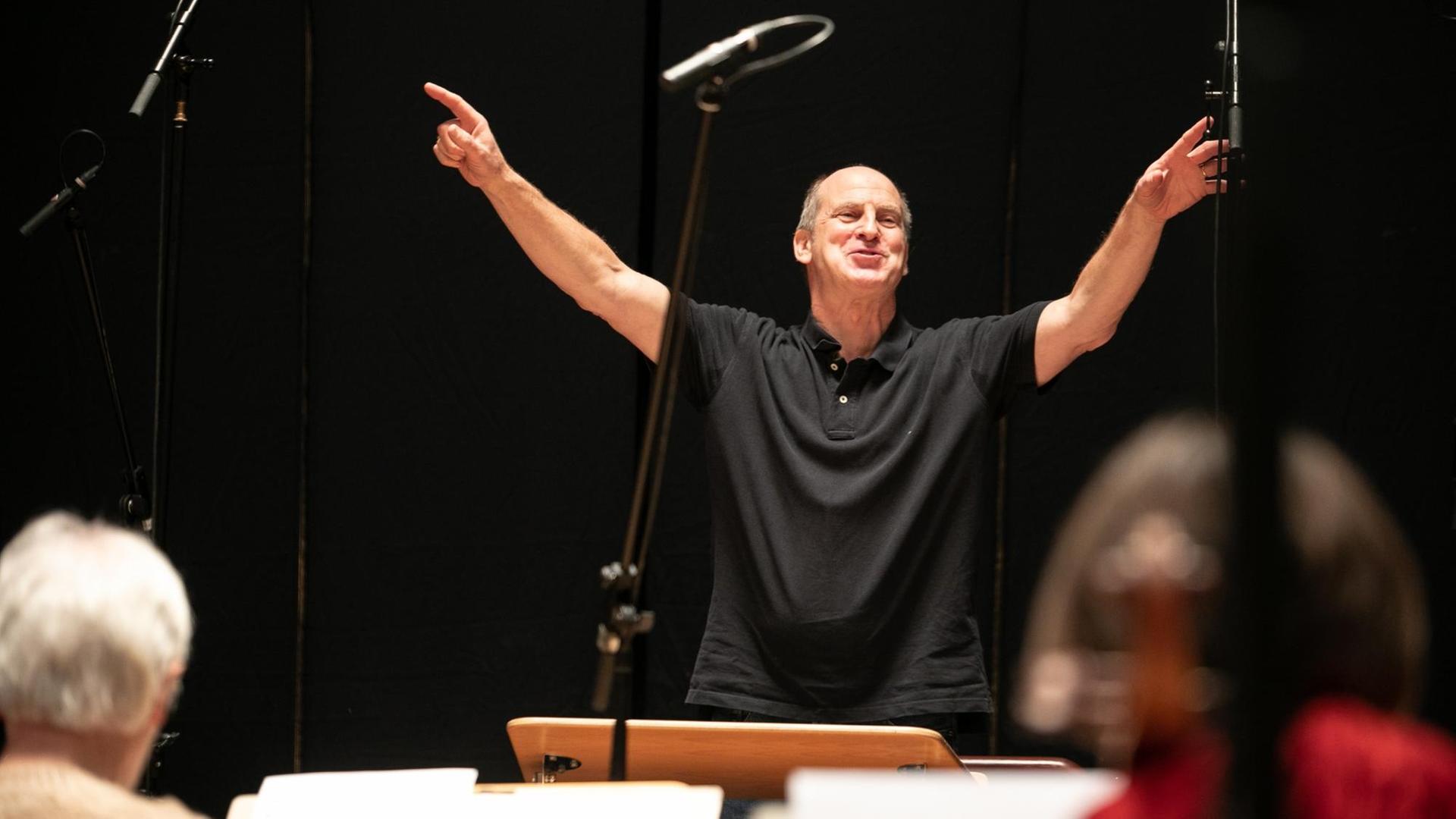 Ein Mann steht vor einem Orchester und reißt die Arme seitlich weit nach oben. Ein Mann steht vor einem Orchester und reißt die Arme seitlich weit nach oben.
