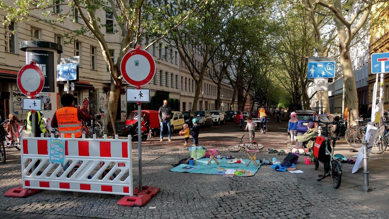 Blick in die Lübbener Straße in Berlin-Kreuzberg. Im Vordergrund die Absperrung für die temporäre Spielstraße, dahinter eine Decke mit Spielzeug, Erwachse und Kinder.