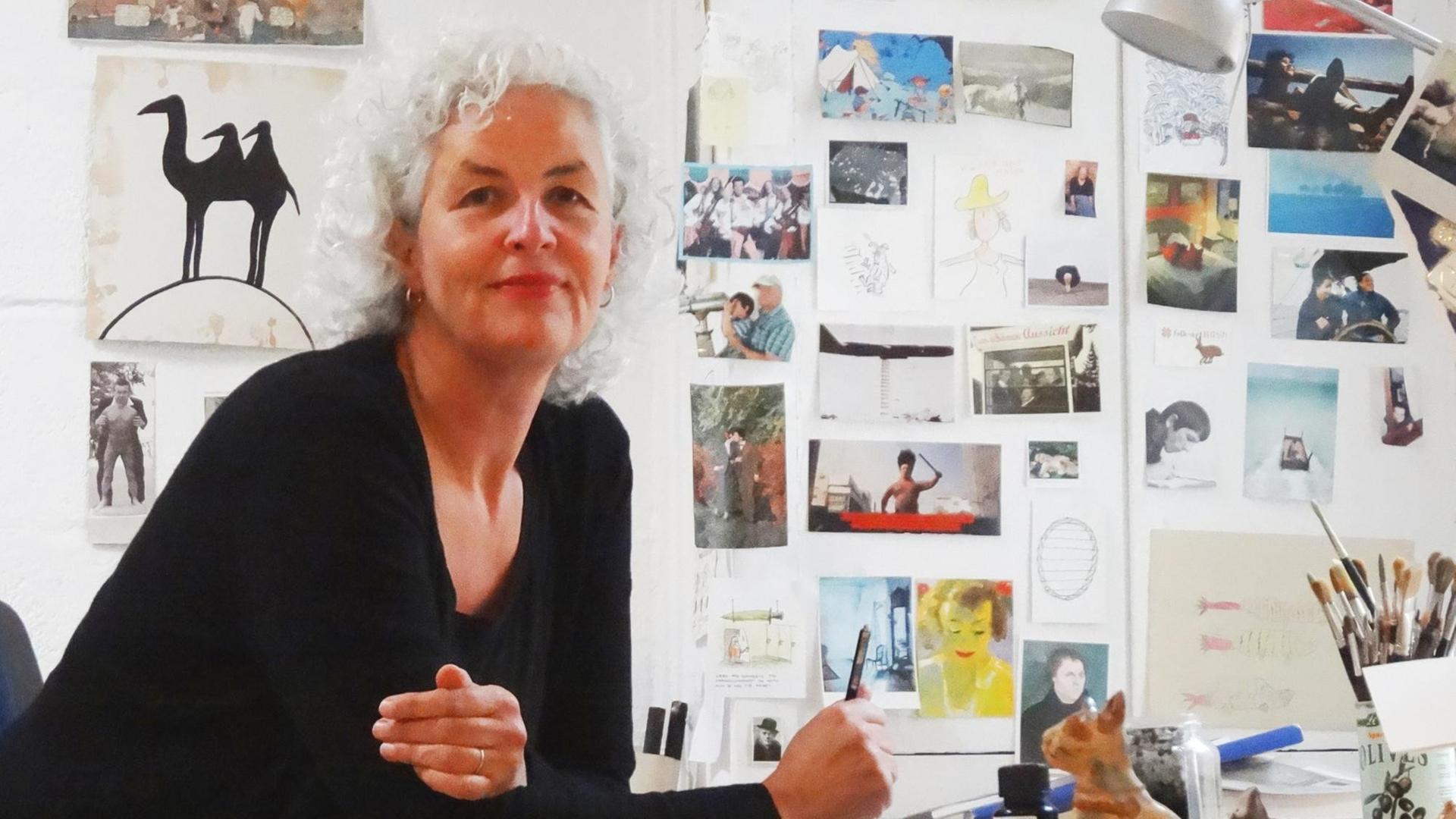 Die Cartoonistin Miriam Wurster sitzt an ihrem Zeichentisch. Um sie herum sind Postkarten, Skizzen und Pinsel zu sehen