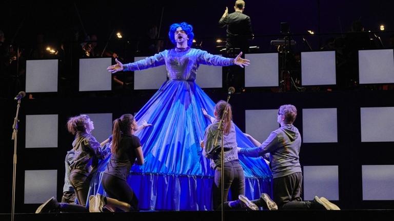 Ein Mann in einem blauen Kleid und einer blauen Perücke hat die Arme ausgebreitet und singt. Vor ihm knien 4 Jugendliche Ein Mann in einem blauen Kleid und einer blauen Perücke hat die Arme ausgebreitet und singt. Vor ihm knien 4 Jugendliche