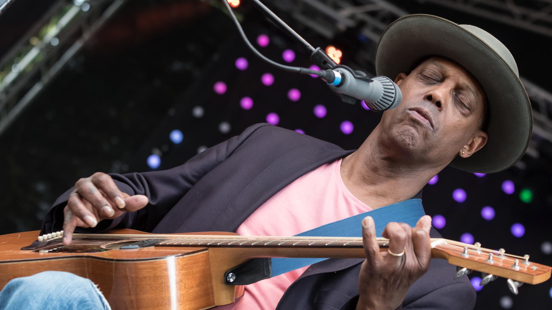 Ein Mann in rosa T-Shirt und mit Hut auf dem Kopf sitzt mit einer Gitarre auf dem Schoß vor einem Mikrofon. Ein Mann in rosa T-Shirt und mit Hut auf dem Kopf sitzt mit einer Gitarre auf dem Schoß vor einem Mikrofon.