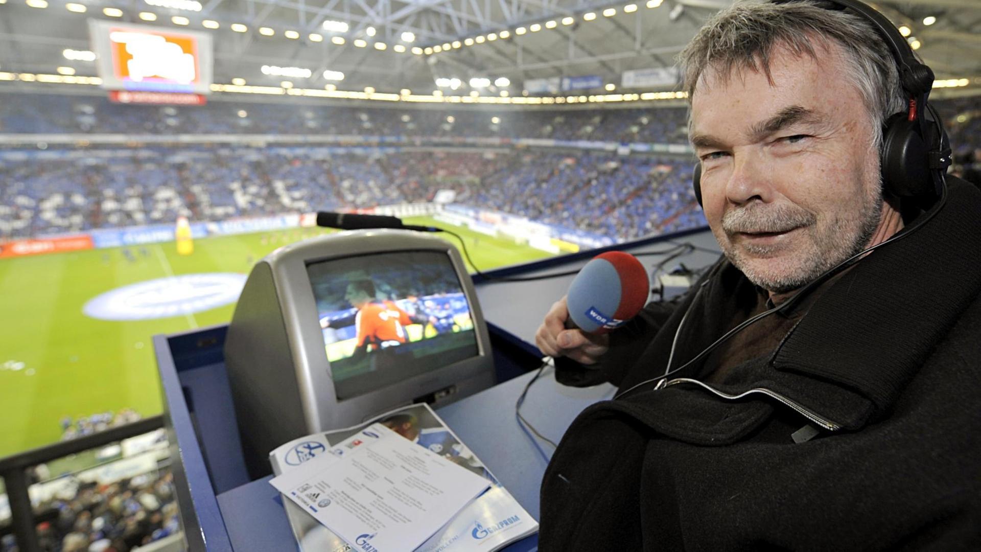Manfred Breuckmann spricht ins Mikrofon, im Hintergrund der Blick auf den Rasen in der Arena Gelsenkirchen.