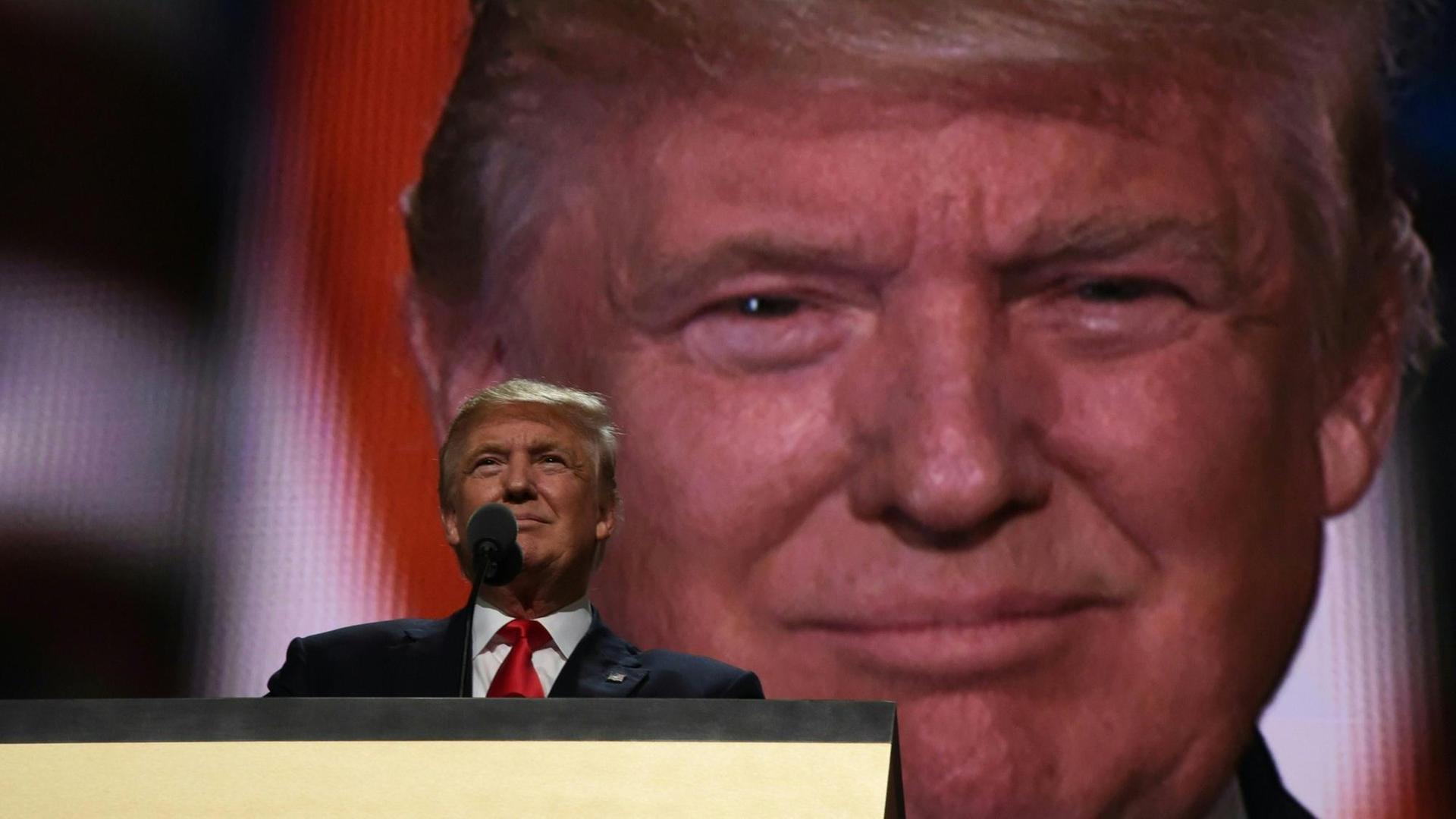 Donald Trump am Rednerpult, hinter ihm sein Gesicht in Großaufnahme auf einer Leinwand