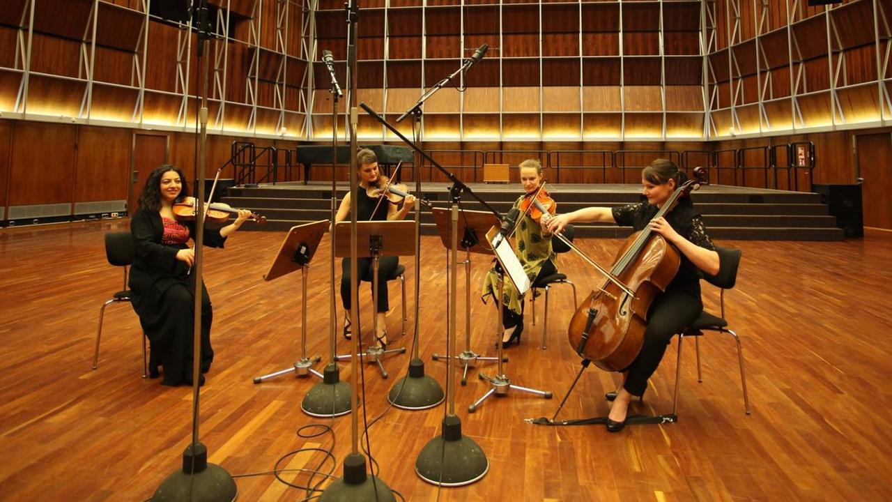 Das Quartett aus vier jungen Musikerinnen sitzt in einem leer geräumten Konzertsaal. Das Quartett aus vier jungen Musikerinnen sitzt in einem leer geräumten Konzertsaal.