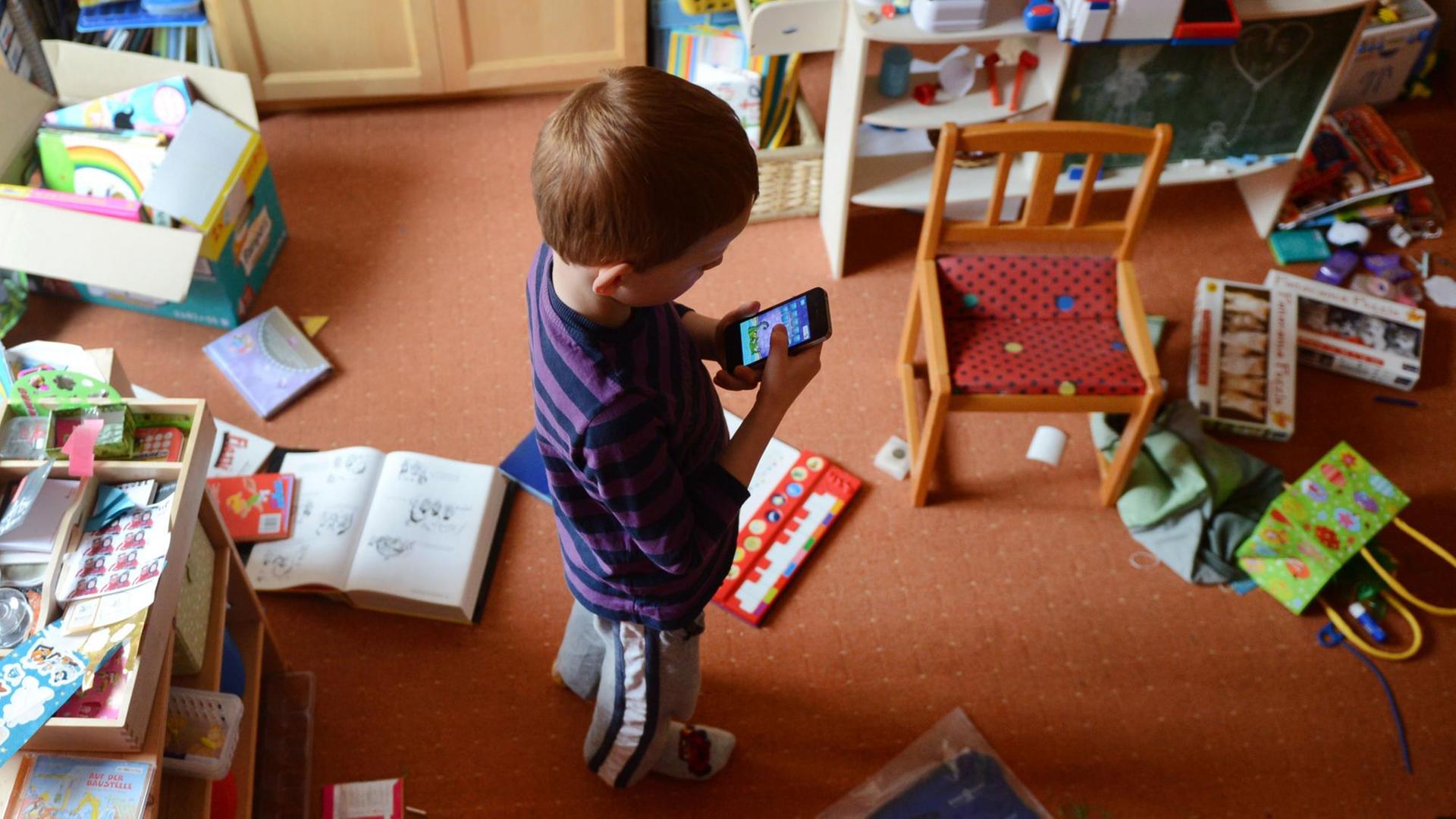 Ein fünfjähriger Junge spielt in seinem unaufgeräumten Kinderzimmer auf einem iPhone ein Computerspiel. Ein fünfjähriger Junge spielt in seinem unaufgeräumten Kinderzimmer auf einem iPhone ein Computerspiel.