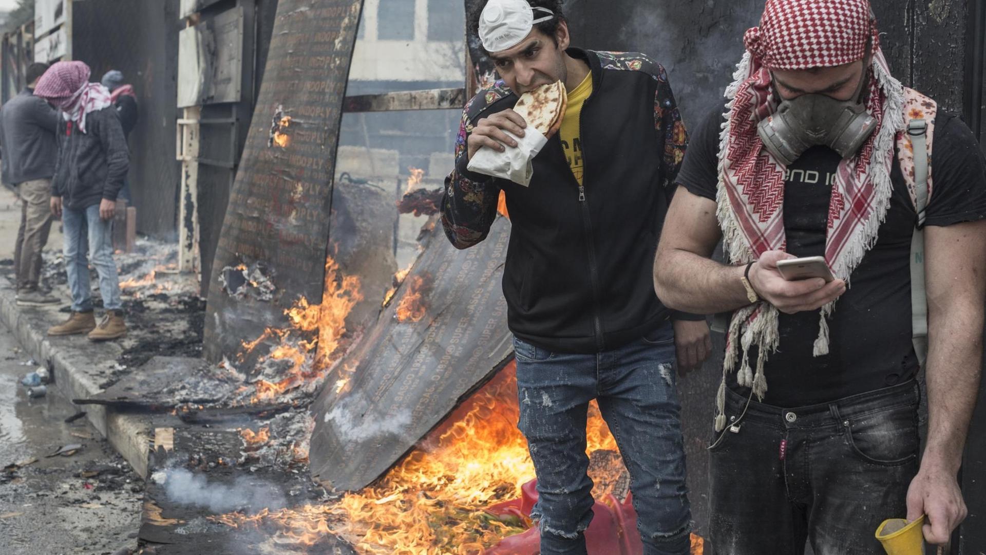 Nach einem gescheiterten Versuch, Politikern den Zugang zum Parlament zu versperren, wo sie über die Annahme der neuen Regierung des Libanon abstimmen werden stärkt sich ein regierungskritischer Demonstrant mit einem traditionellen libanesischen Sandwich, ein zweiter mit Gasmaske und Palästinenser-Tuch schaut auf sein Handy. Im Hintergrund brennt ein Bauzaun. Nach einem gescheiterten Versuch, Politikern den Zugang zum Parlament zu versperren, wo sie über die Annahme der neuen Regierung des Libanon abstimmen werden stärkt sich ein regierungskritischer Demonstrant mit einem traditionellen libanesischen Sandwich, ein zweiter mit Gasmaske und Palästinenser-Tuch schaut auf sein Handy. Im Hintergrund brennt ein Bauzaun.