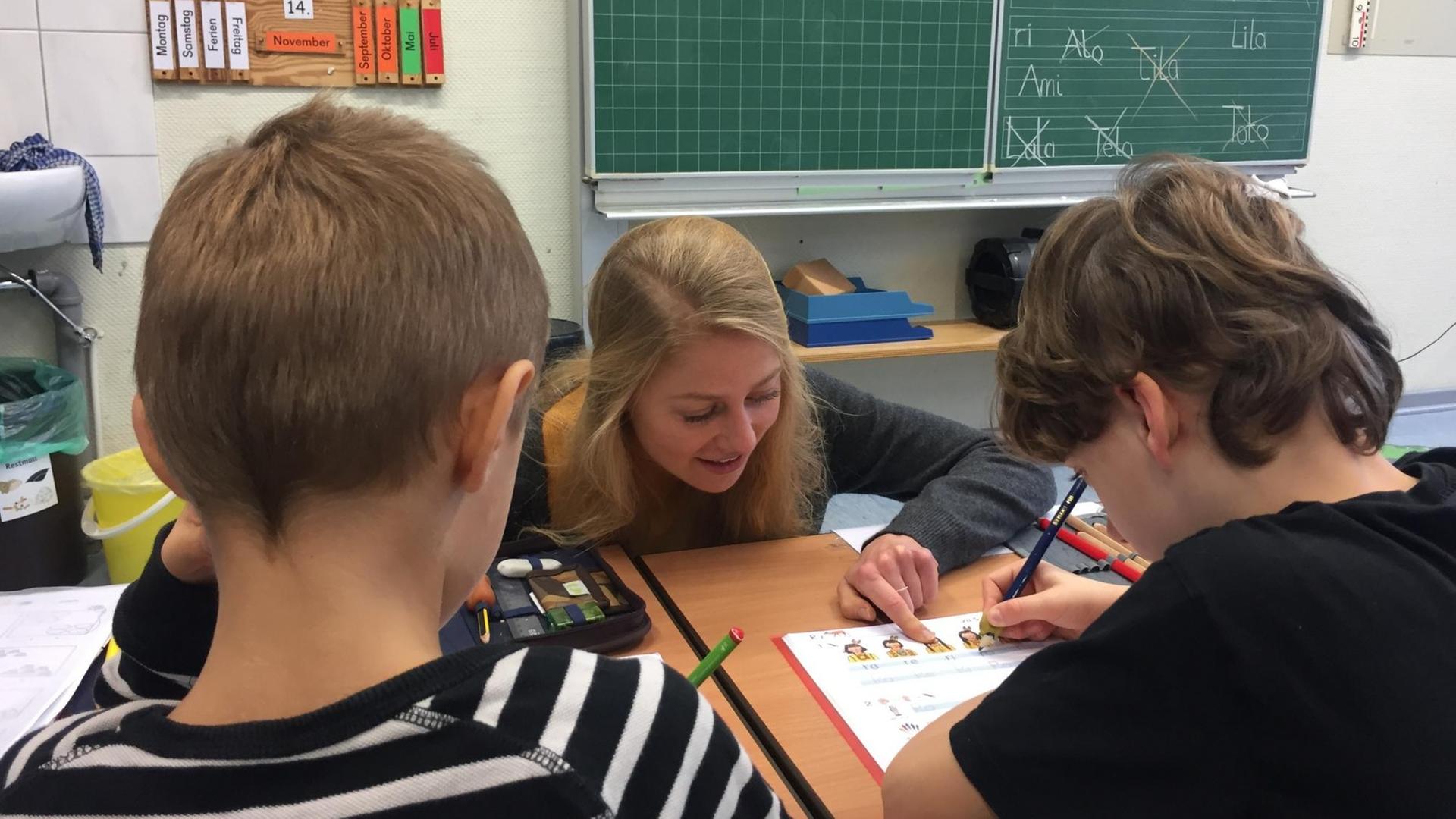 Luzie Niedworok hockt vor einem Schreibtisch von zwei Erstklässlern und erklärt ihnen eine Aufgabe. Die Masterstudentin Niedworok hat gemeinsam mit 13 weiteren Studierenden für eine Woche lang die Grundschule Jübek / Schleswig-Holstein adoptiert und meistert den Schulalltag allein. | Foto: Birgitta von Gyldenfeldt/dpa | Verwendung weltweit Luzie Niedworok hockt vor einem Schreibtisch von zwei Erstklässlern und erklärt ihnen eine Aufgabe. Die Masterstudentin Niedworok hat gemeinsam mit 13 weiteren Studierenden für eine Woche lang die Grundschule Jübek / Schleswig-Holstein adoptiert und meistert den Schulalltag allein. | Foto: Birgitta von Gyldenfeldt/dpa | Verwendung weltweit