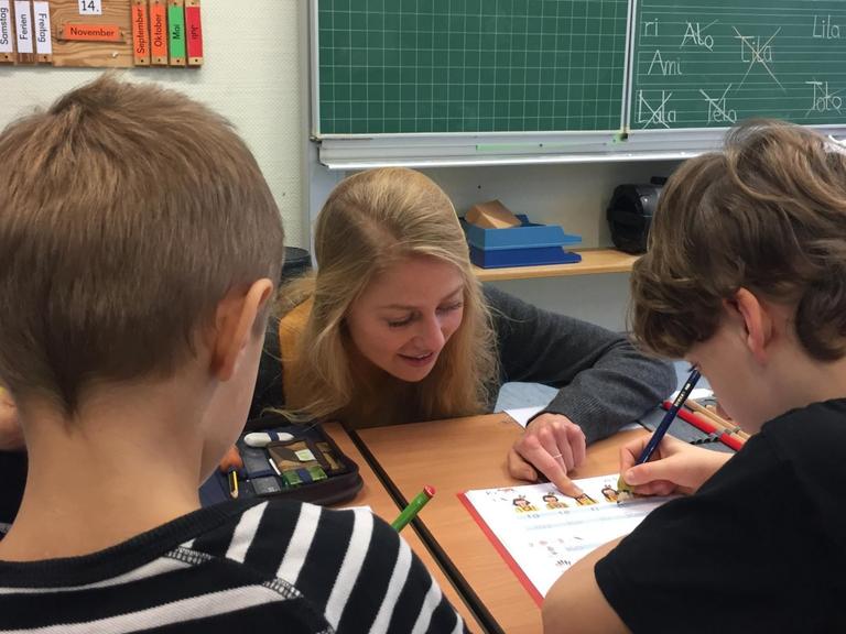 Luzie Niedworok hockt vor einem Schreibtisch von zwei Erstklässlern und erklärt ihnen eine Aufgabe. Die Masterstudentin Niedworok hat gemeinsam mit 13 weiteren Studierenden für eine Woche lang die Grundschule Jübek / Schleswig-Holstein adoptiert und meistert den Schulalltag allein. | Foto: Birgitta von Gyldenfeldt/dpa | Verwendung weltweit
