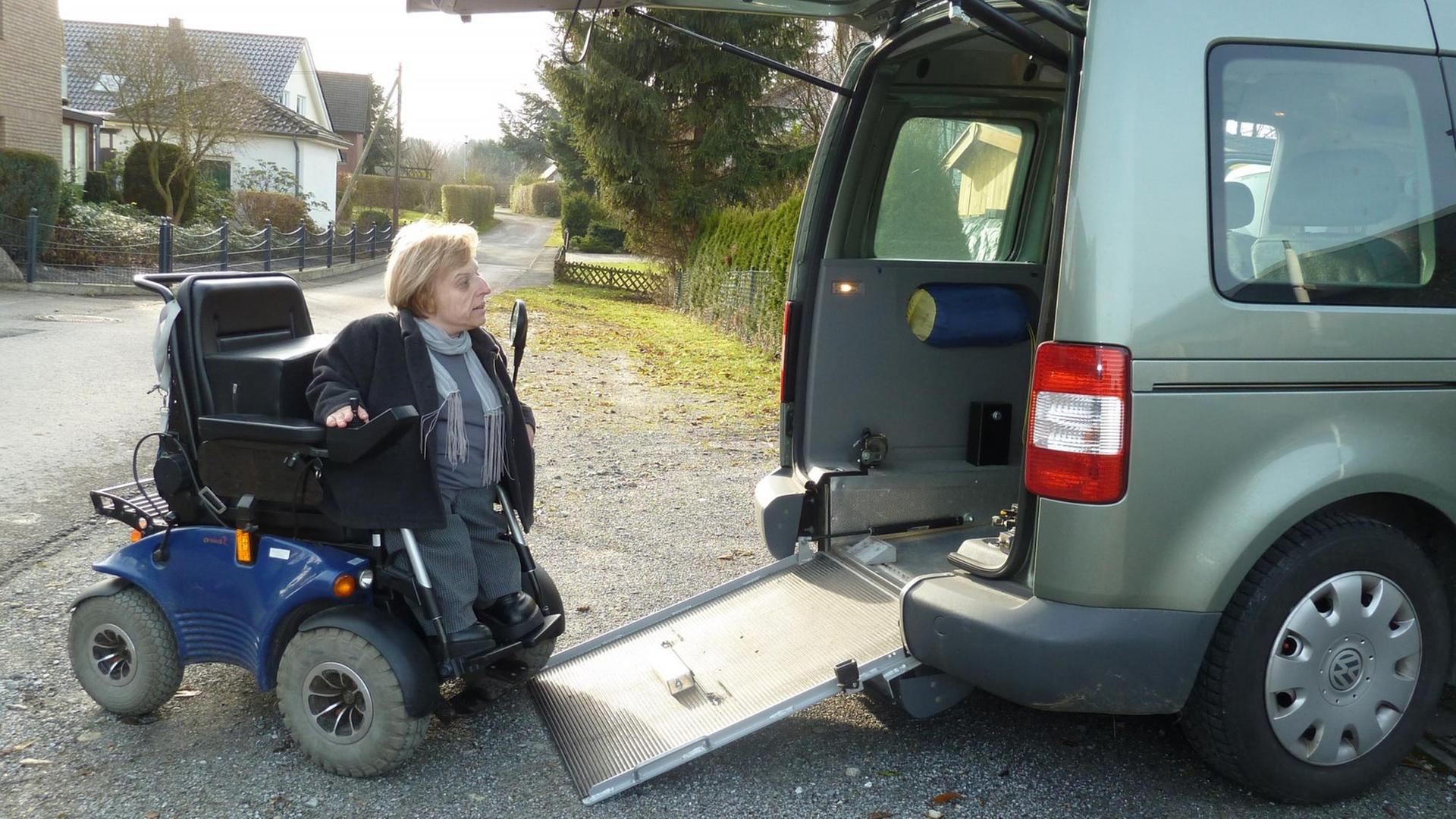 Eine Frau sitzt in einem Rollstuhl und fährt auf einer kleinen Rampe in den hinteren Bereich eines größeren Autos. Eine Frau sitzt in einem Rollstuhl und fährt auf einer kleinen Rampe in den hinteren Bereich eines größeren Autos.