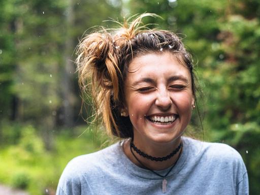Ein junge Frau steht auf einem Weg im Wald und lacht mit geschlossenen Augen.