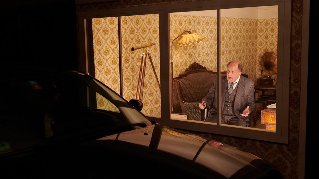 Es ist dunkel. Durch ein großes Fenster fällt der Blick in ein erhelltes Wohnzimmer, in dem ein Mann im Sessel sitzt und nach draußen schaut. Es ist dunkel. Durch ein großes Fenster fällt der Blick in ein erhelltes Wohnzimmer, in dem ein Mann im Sessel sitzt und nach draußen schaut.