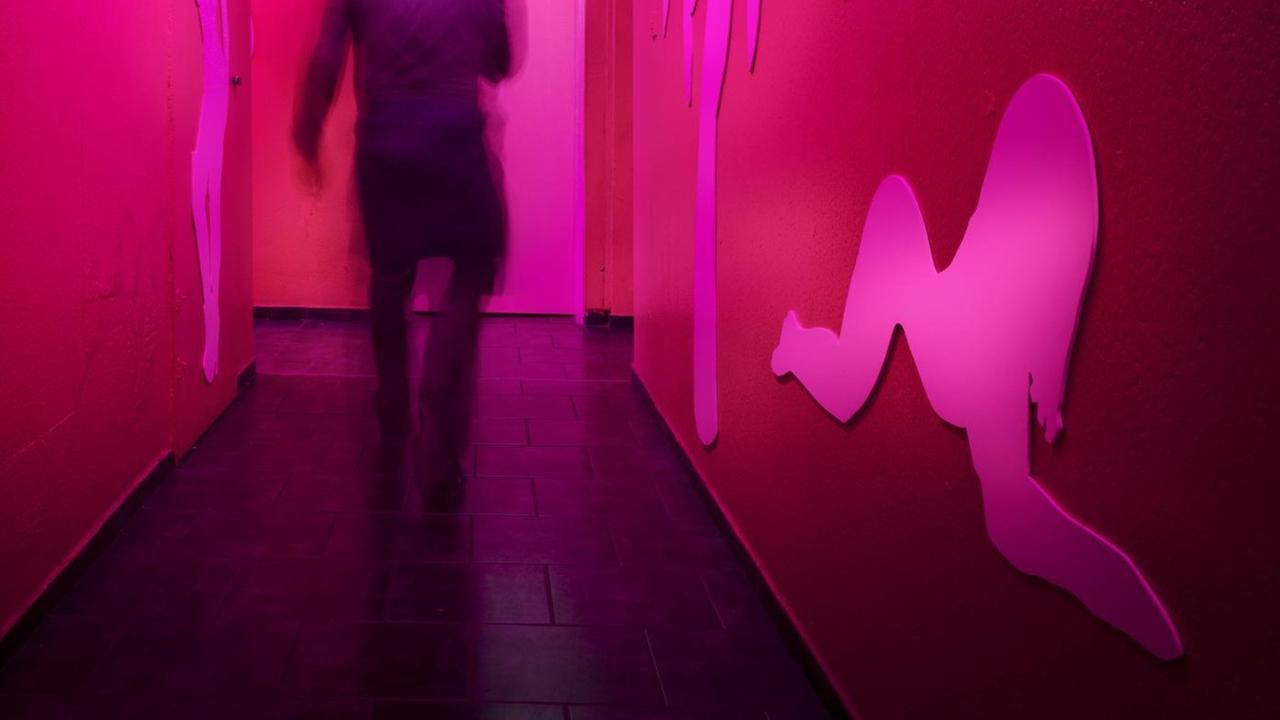 Der Rücken einer Person ist in Bewegungsunschärfe auf einem rot beleuchteten Flur eines Bordells zu sehen. An den Wänden ist die Silhouette einer Fau zu erkennen. Der Rücken einer Person ist in Bewegungsunschärfe auf einem rot beleuchteten Flur eines Bordells zu sehen. An den Wänden ist die Silhouette einer Fau zu erkennen.