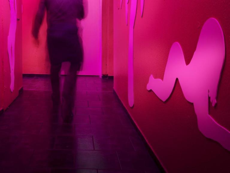 Der Rücken einer Person ist in Bewegungsunschärfe auf einem rot beleuchteten Flur eines Bordells zu sehen. An den Wänden ist die Silhouette einer Fau zu erkennen.