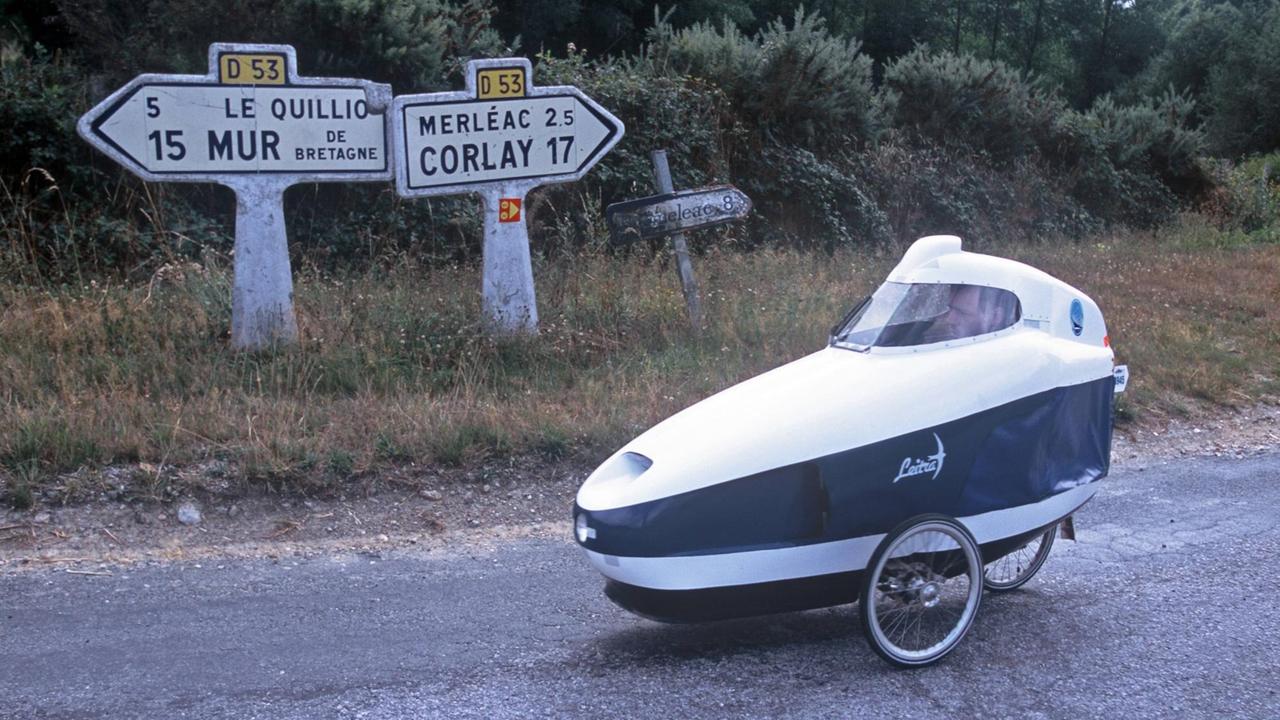 Ein Teilnehmer in seinem Leitra Velomobil auf einer Landstrasse, im Hintergund französische Strassenschilder. Ein Teilnehmer in seinem Leitra Velomobil auf einer Landstrasse, im Hintergund französische Strassenschilder.