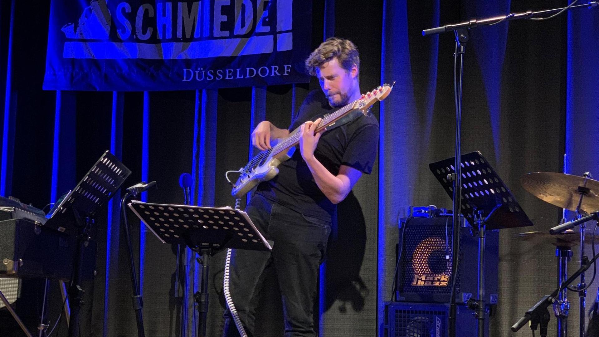 Ein junger Mann spielt in blaues Licht gehüllt auf einer Bühne E-Bass und blickt konzentriert auf ein Notenpult. Der Oberkörper ist elastisch zurückgebeugt.