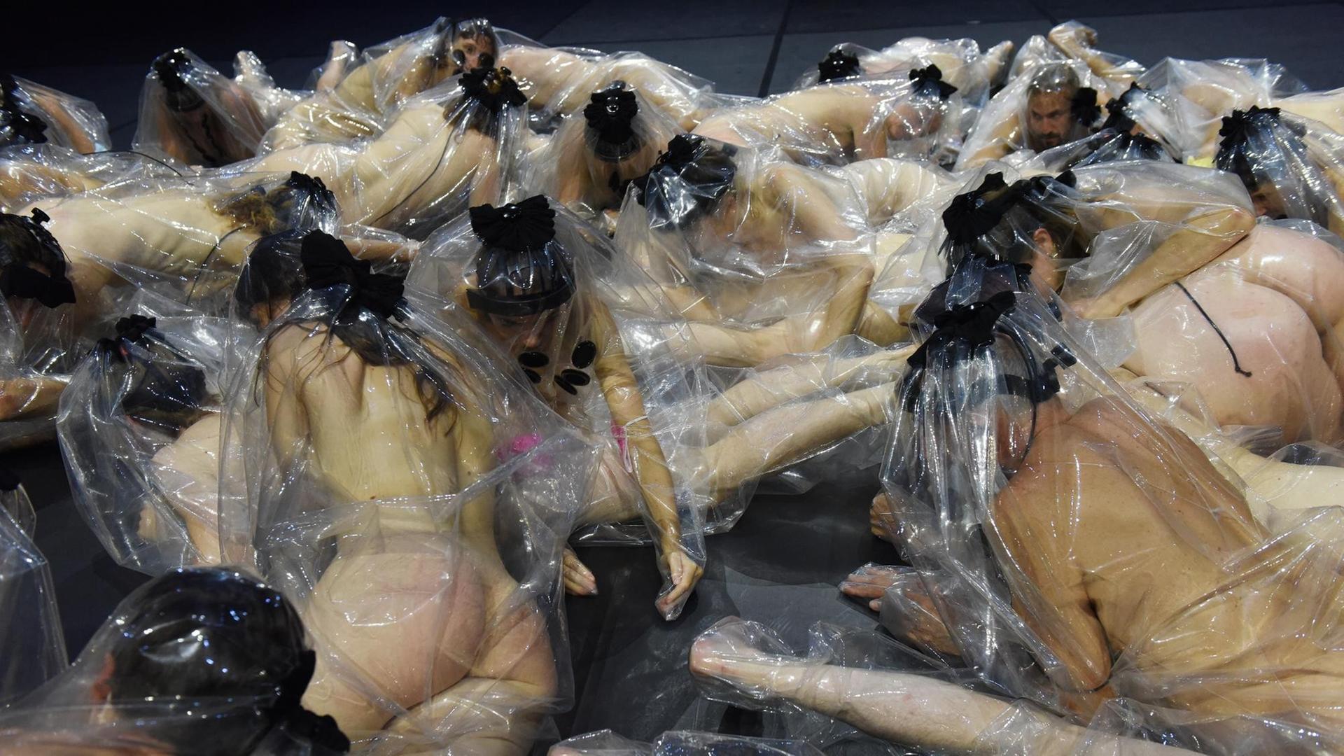 Nackte Männer und Frauen, in transparente Folien gehüllt, bewegen sich nebeneinander auf einem schwarzen Bühnenboden. Nackte Männer und Frauen, in transparente Folien gehüllt, bewegen sich nebeneinander auf einem schwarzen Bühnenboden.