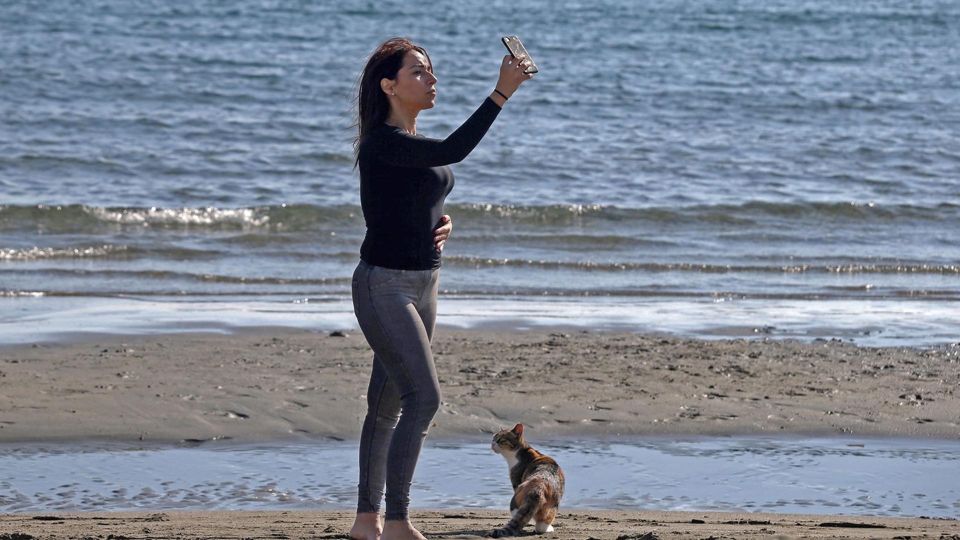 Eine Frau schießt am Strand von Limassol (Zypern) ein Selfie. Zu ihren Füßen sitzt ein braungescheckte Katze. Eine Frau schießt am Strand von Limassol (Zypern) ein Selfie. Zu ihren Füßen sitzt ein braungescheckte Katze.