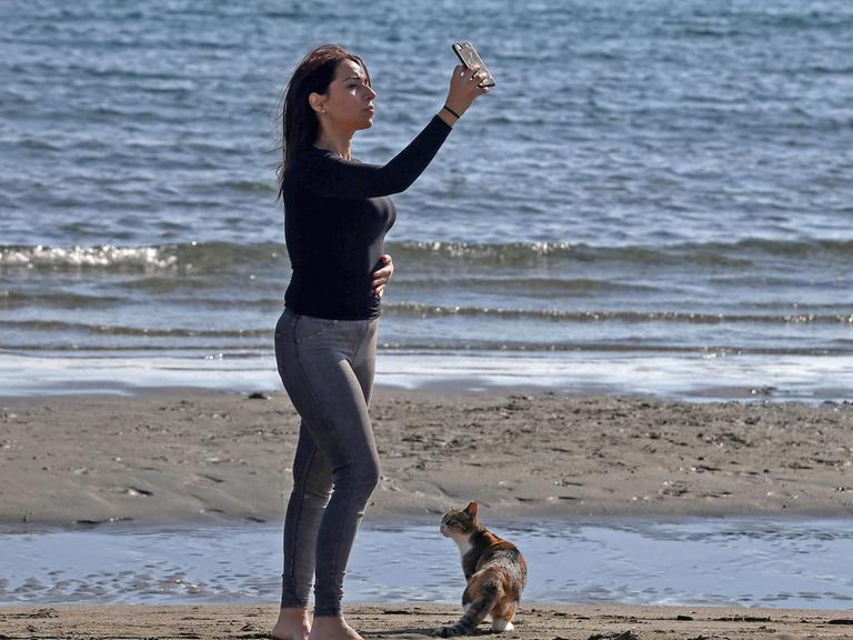 Eine Frau schießt am Strand von Limassol (Zypern) ein Selfie. Zu ihren Füßen sitzt ein braungescheckte Katze.