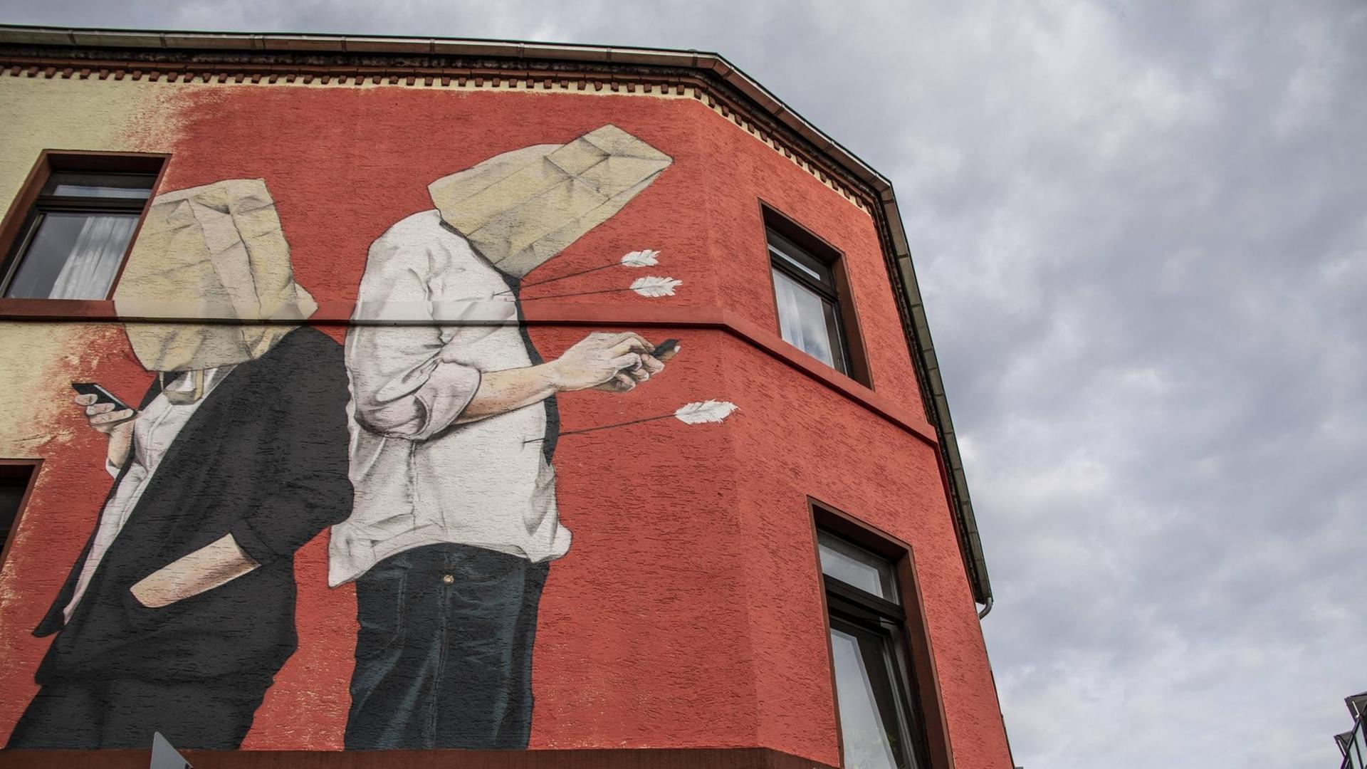 Ein Bild an einer Hauswand: Zwei Menschen, die Tüten über dem Kopf und Handys in der Hand haben. Ein Bild an einer Hauswand: Zwei Menschen, die Tüten über dem Kopf und Handys in der Hand haben.