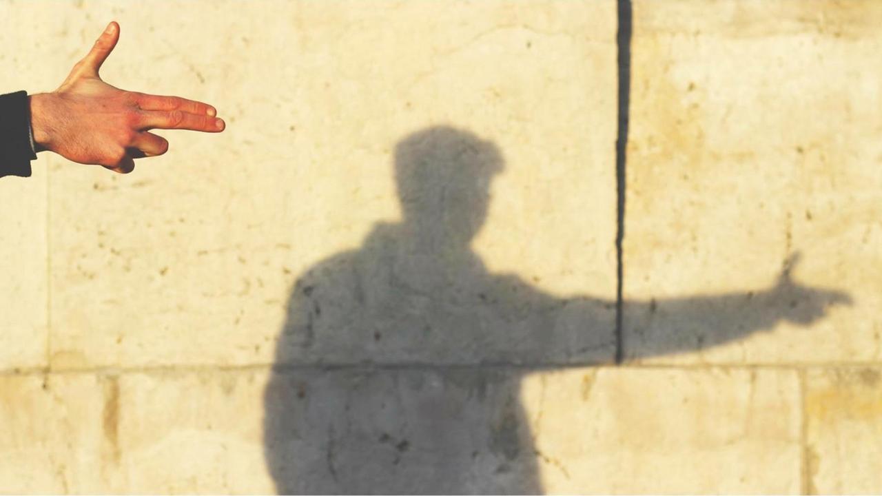 An der Wand sieht man den Schatten eines Mannes, der mit der Hand eine Pistole formt. An der Wand sieht man den Schatten eines Mannes, der mit der Hand eine Pistole formt.