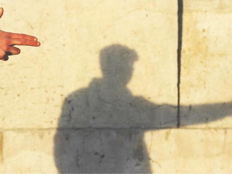 An der Wand sieht man den Schatten eines Mannes, der mit der Hand eine Pistole formt.