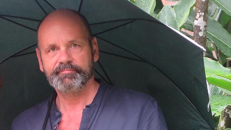 Ein Mann mit Vollbart und dunklem Schirm steht vor einer Grünpflanze. Ein Mann mit Vollbart und dunklem Schirm steht vor einer Grünpflanze.