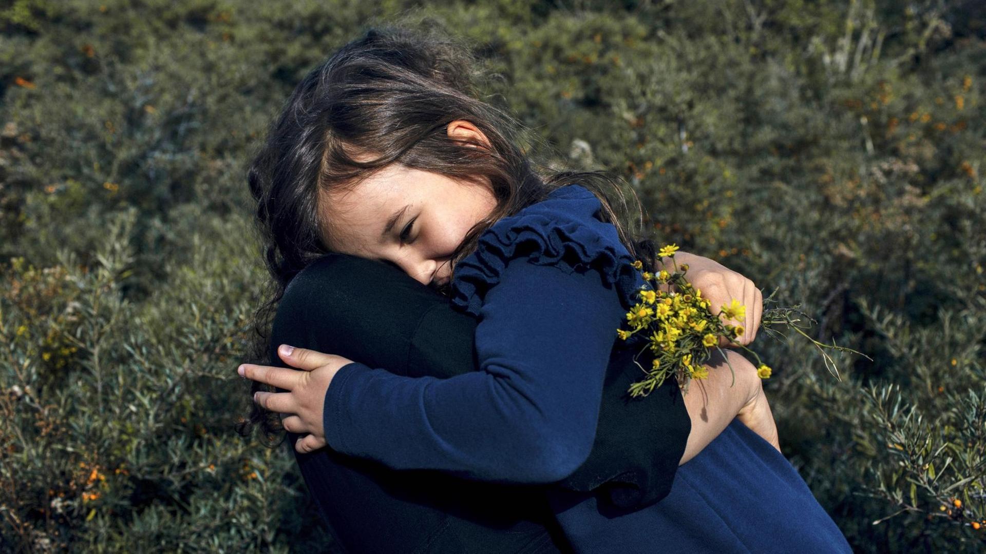 Ein Mädchen in einem blauen Kleid umarmt eine Frau, welche goldgelbe Blumen in der Hand hat. Ein Mädchen in einem blauen Kleid umarmt eine Frau, welche goldgelbe Blumen in der Hand hat.