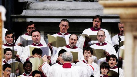 Ein Chor aus Knaben und Männern in langen weißen Kutten und dunkelroten Krägen singt in einem Kirchenraum.