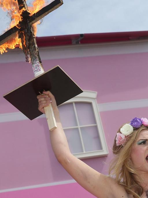 Eine Femen-Demonstrantin mit Blumen im Haar und nackten Brüsten protestiert vor dem Barbie House der Firma Mattel in Berlin, sie hält mit der rechten Hand ein brennendes Kreuz in die Höhe, an das eine Barbie-Puppe gebunden ist, und hat auf ihren Oberkörper die Worte geschrieben: "A Life In Plastic Is Not Fantastic" (Ein Plastik-Leben ist nicht fantastisch).
