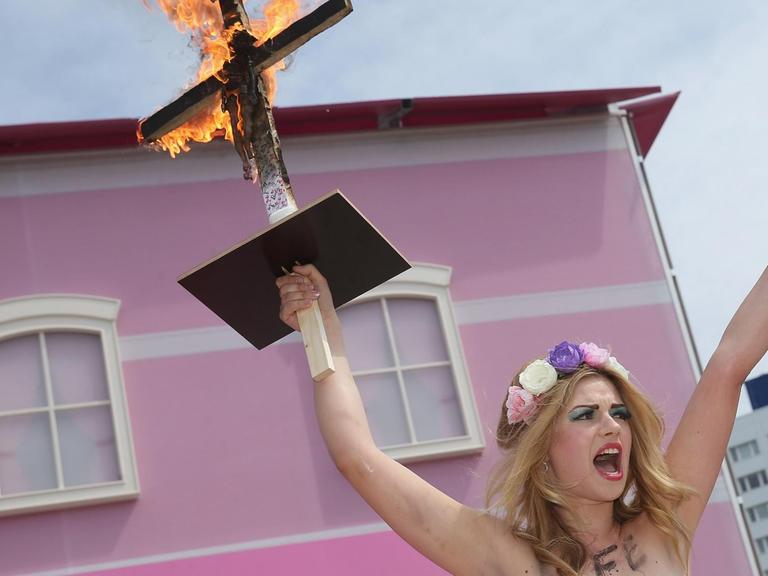 Eine Femen-Demonstrantin mit Blumen im Haar und nackten Brüsten protestiert vor dem Barbie House der Firma Mattel in Berlin, sie hält mit der rechten Hand ein brennendes Kreuz in die Höhe, an das eine Barbie-Puppe gebunden ist, und hat auf ihren Oberkörper die Worte geschrieben: "A Life In Plastic Is Not Fantastic" (Ein Plastik-Leben ist nicht fantastisch).