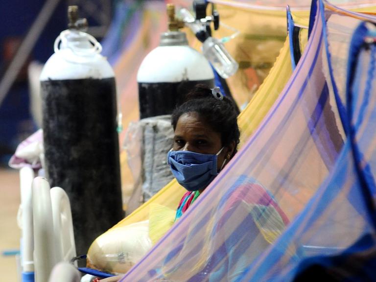 Eine Patientin sitzt auf einem Bett, sie trägt einen Mundnasenschutz. Hinter ihr ist eine Sauerstoff-Flasche zu sehen.