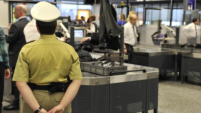 Eine Polizistin in grüner Uniform steht mit hinter dem Rücken verschränkten Armen (und dem Rücken zu Kamera) vor zwei Laufbändern, auf denen Gepäck von Passagieren kontrolliert wird.