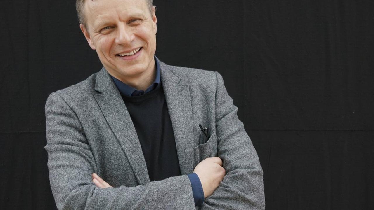 Der Autor und Jurist steht vor einer schwarzen Wand, trägt ein graues Sacko und lacht in die Kamera. Der Autor und Jurist steht vor einer schwarzen Wand, trägt ein graues Sacko und lacht in die Kamera.