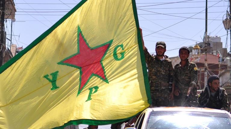 Männer halten aus einem Auto heraus eine Flagge YPG-Miliz