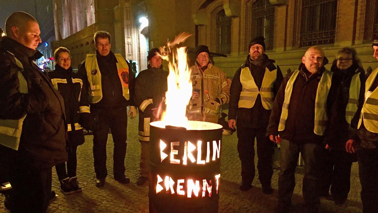 Neun Menschen stehen um eine Feuertonne, es ist dunkel. In die Tonne sind die Worte "Berlin brennt" eingestanzt, die nun durch die Tonne leuchten. Neun Menschen stehen um eine Feuertonne, es ist dunkel. In die Tonne sind die Worte "Berlin brennt" eingestanzt, die nun durch die Tonne leuchten.