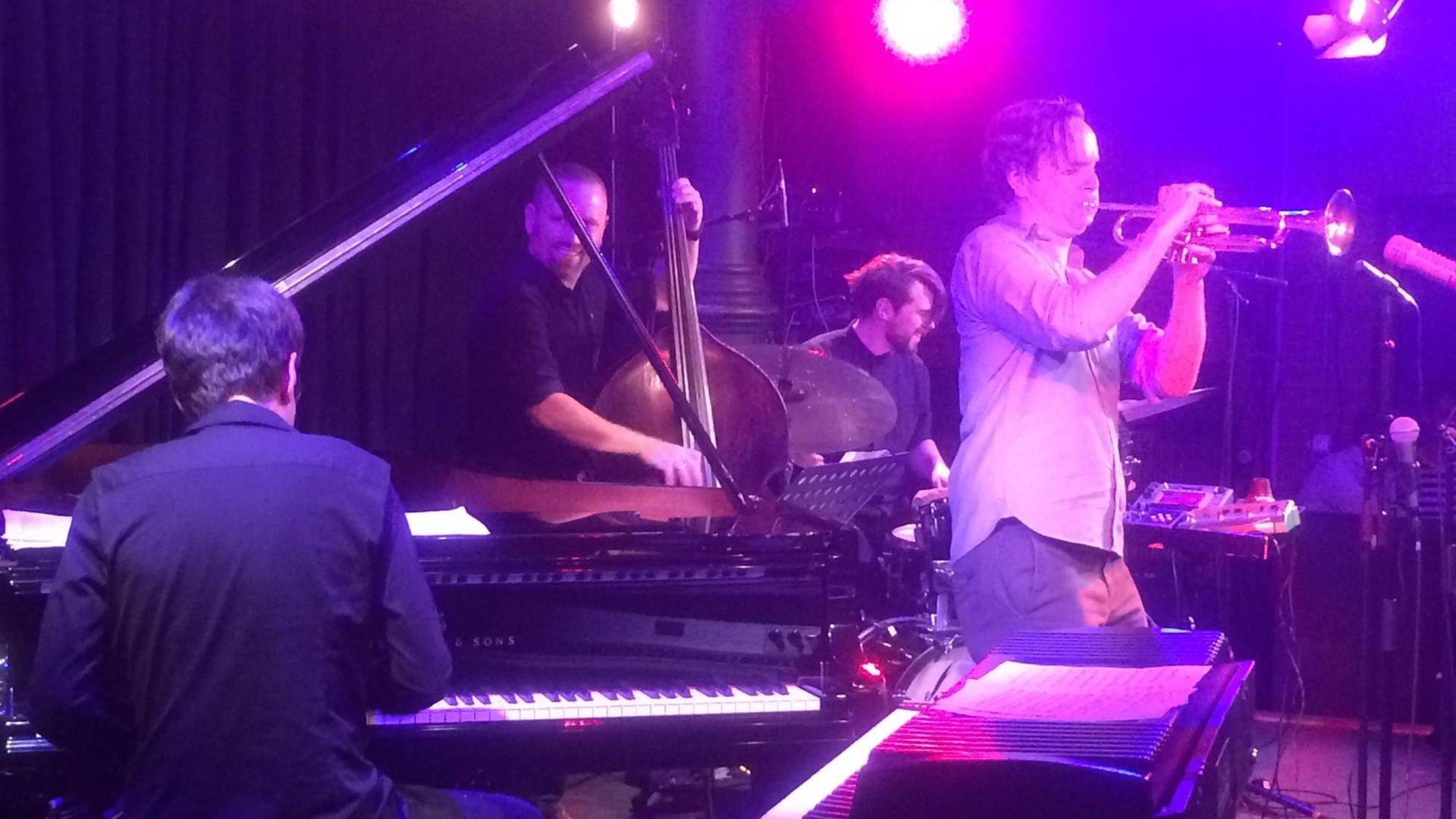 Ein Konzertbild in violett-rotem Bühnenlicht. Vier Musiker sind auf der Bühne versammelt, links der Pianist nur rückseitig zu sehen, in der Mitte hinten der Kontrabassist und der Schlagzeuger. Rechts bläst der Trompeter in sein Instrument. Ein Konzertbild in violett-rotem Bühnenlicht. Vier Musiker sind auf der Bühne versammelt, links der Pianist nur rückseitig zu sehen, in der Mitte hinten der Kontrabassist und der Schlagzeuger. Rechts bläst der Trompeter in sein Instrument.