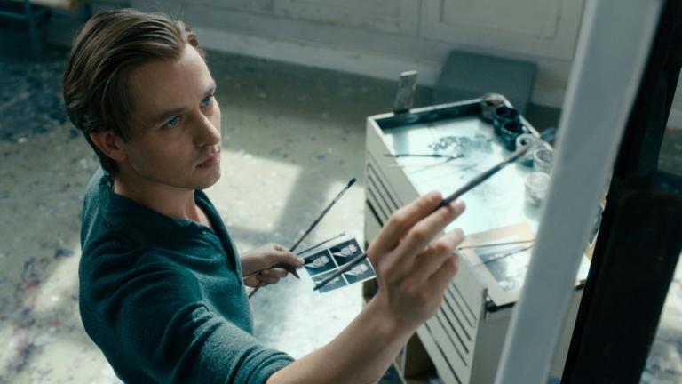 Ein junger Künstler (Tom Schilling) mit einem Foto in der einen und einem Pinsel in der anderen Hand vor einer Leinwand Ein junger Künstler (Tom Schilling) mit einem Foto in der einen und einem Pinsel in der anderen Hand vor einer Leinwand