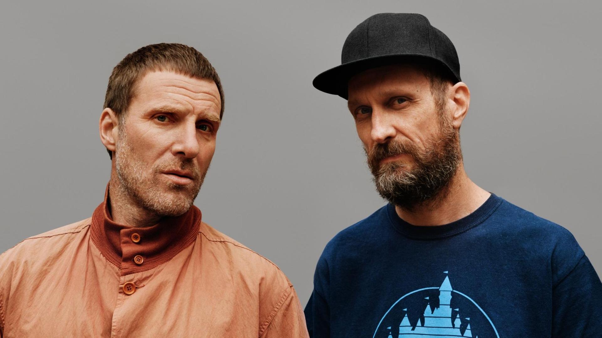 Zwei Männer blicken in die Kamera. Der linke Mann hat kurze Haare und trägt eine helle Jacke. Der rechte Mann trägt ein T-Shirt und eine Baseball-Kappe. Zwei Männer blicken in die Kamera. Der linke Mann hat kurze Haare und trägt eine helle Jacke. Der rechte Mann trägt ein T-Shirt und eine Baseball-Kappe.