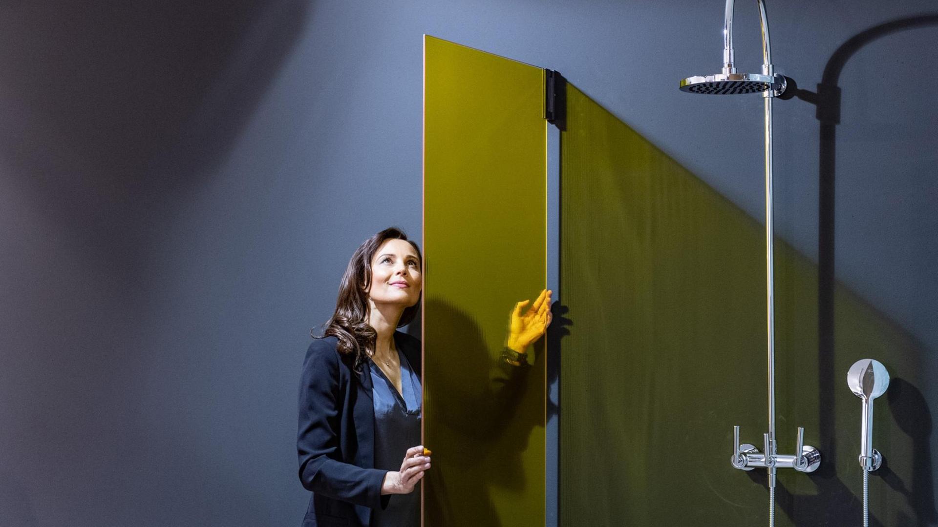 Ein Frau steht auf der Messe in einem arrangierten Badezimmer mit grauer Wand und einer gelben Glasduschtür. Ein Frau steht auf der Messe in einem arrangierten Badezimmer mit grauer Wand und einer gelben Glasduschtür.