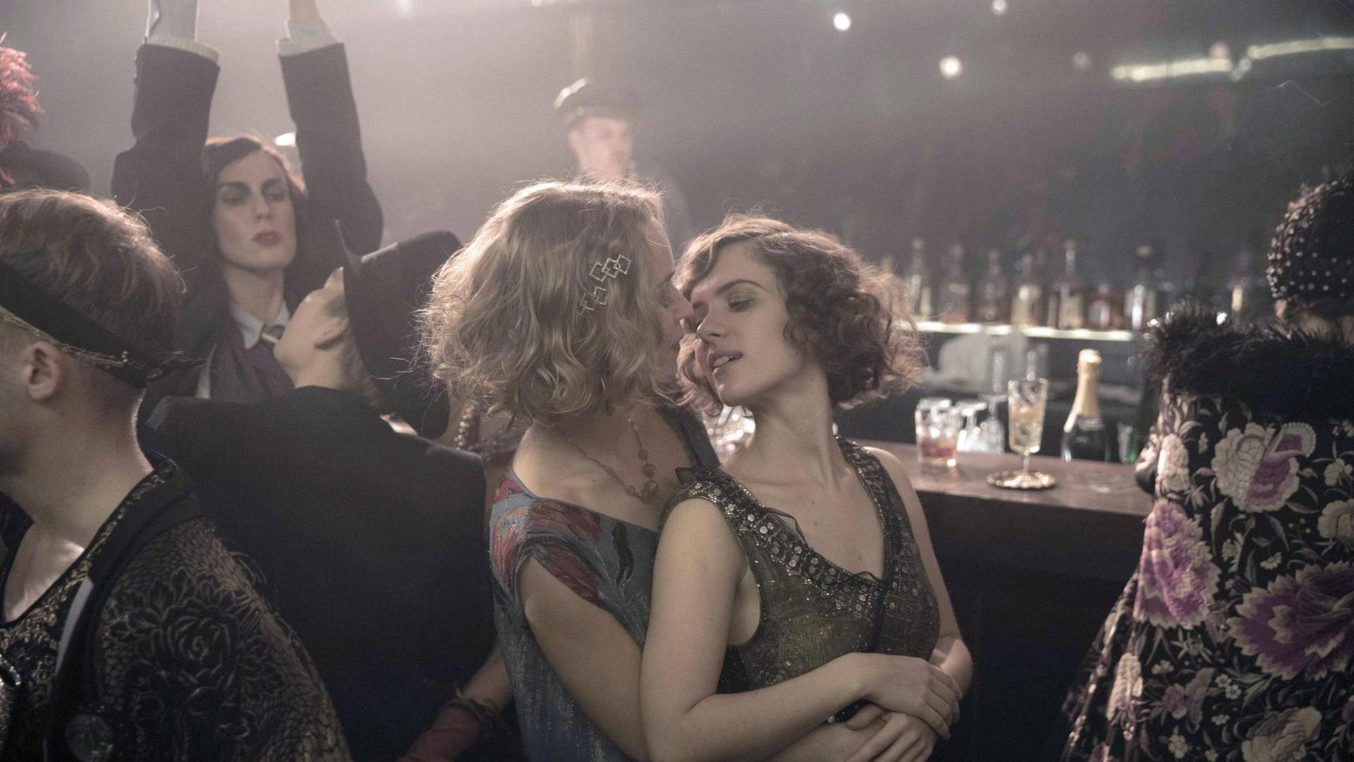 Partyszene aus der TV-Serie "Babylon Berlin" im Stil der 20er-Jahre: Eine Frau umarmt eine andere von hinten, ihre Gesichter sind einander zugewandt, darum mehrere Menschen, manche an einem Tresen, eine Frau reckt beide Arme in die Höhe. Partyszene aus der TV-Serie "Babylon Berlin" im Stil der 20er-Jahre: Eine Frau umarmt eine andere von hinten, ihre Gesichter sind einander zugewandt, darum mehrere Menschen, manche an einem Tresen, eine Frau reckt beide Arme in die Höhe.