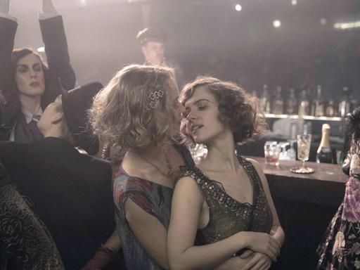 Partyszene aus der TV-Serie "Babylon Berlin" im Stil der 20er-Jahre: Eine Frau umarmt eine andere von hinten, ihre Gesichter sind einander zugewandt, darum mehrere Menschen, manche an einem Tresen, eine Frau reckt beide Arme in die Höhe.