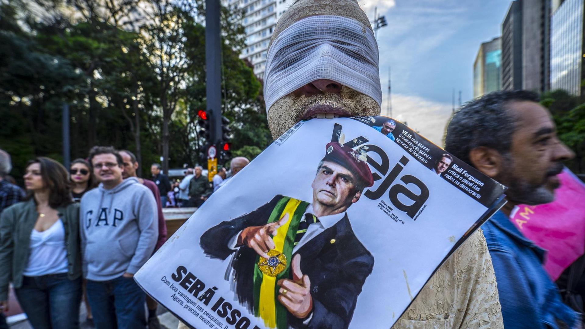 Ein verkleideter Demonstrant mit Binde über der Augenpartie und einer Zeitung, die Jair Bolsonaro abbildet, im Mund, nimmt an Protesten gegen den neuen Präsidenten Brasiliens teil.