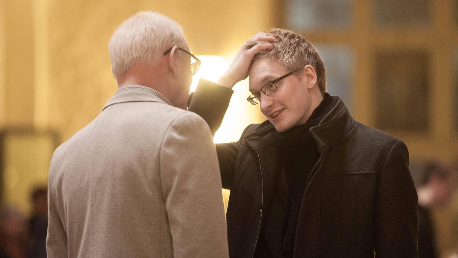 Farbfoto eines jungen blonden Mannes, der sich im Gespräch mit einem älteren Mann befindet, den man nur von hinten sieht Farbfoto eines jungen blonden Mannes, der sich im Gespräch mit einem älteren Mann befindet, den man nur von hinten sieht